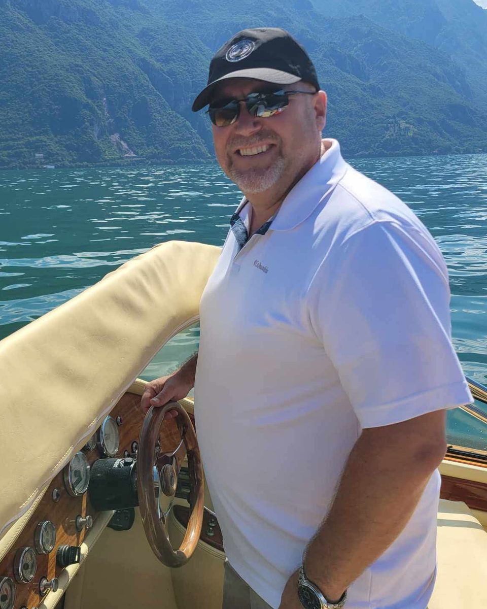 Monty smiling at the wheel of a wooden boat on a mountain lake