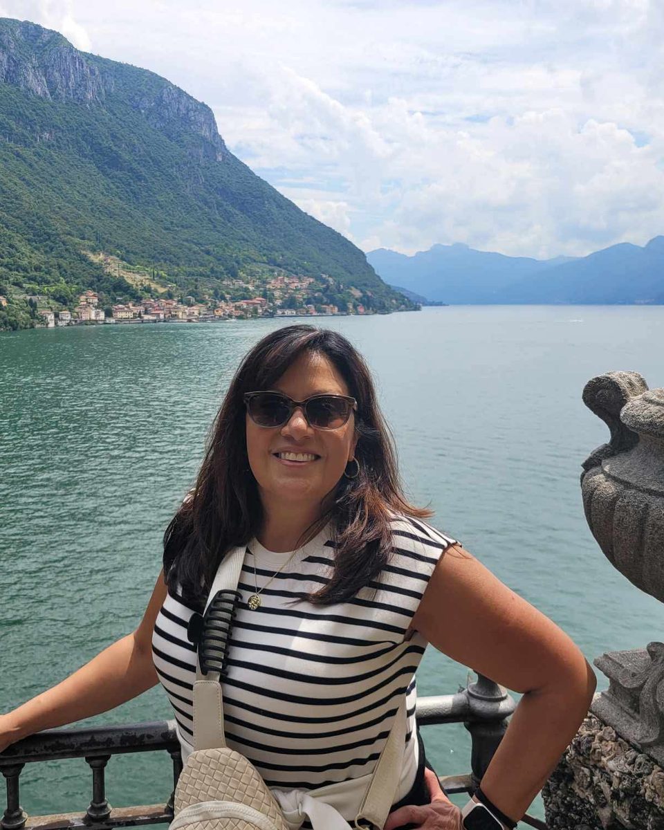 Nita smiling on a terrace above a lake, with green mountains behind her